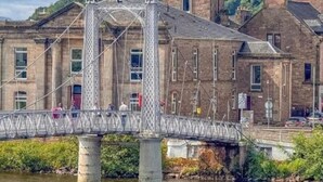 Un puente con estructura metálica y pilares de piedra sobre un río, con edificios y una torre al fondo.