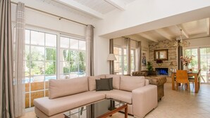 Amplia sala de estar con sofá seccional color beige, una mesa de centro de vidrio y una chimenea. Amplias ventanas ofrecen vistas al exterior.