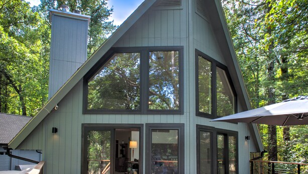 Una cabaña moderna y de tonos claros con ventanas amplias y una terraza, rodeada de árboles.