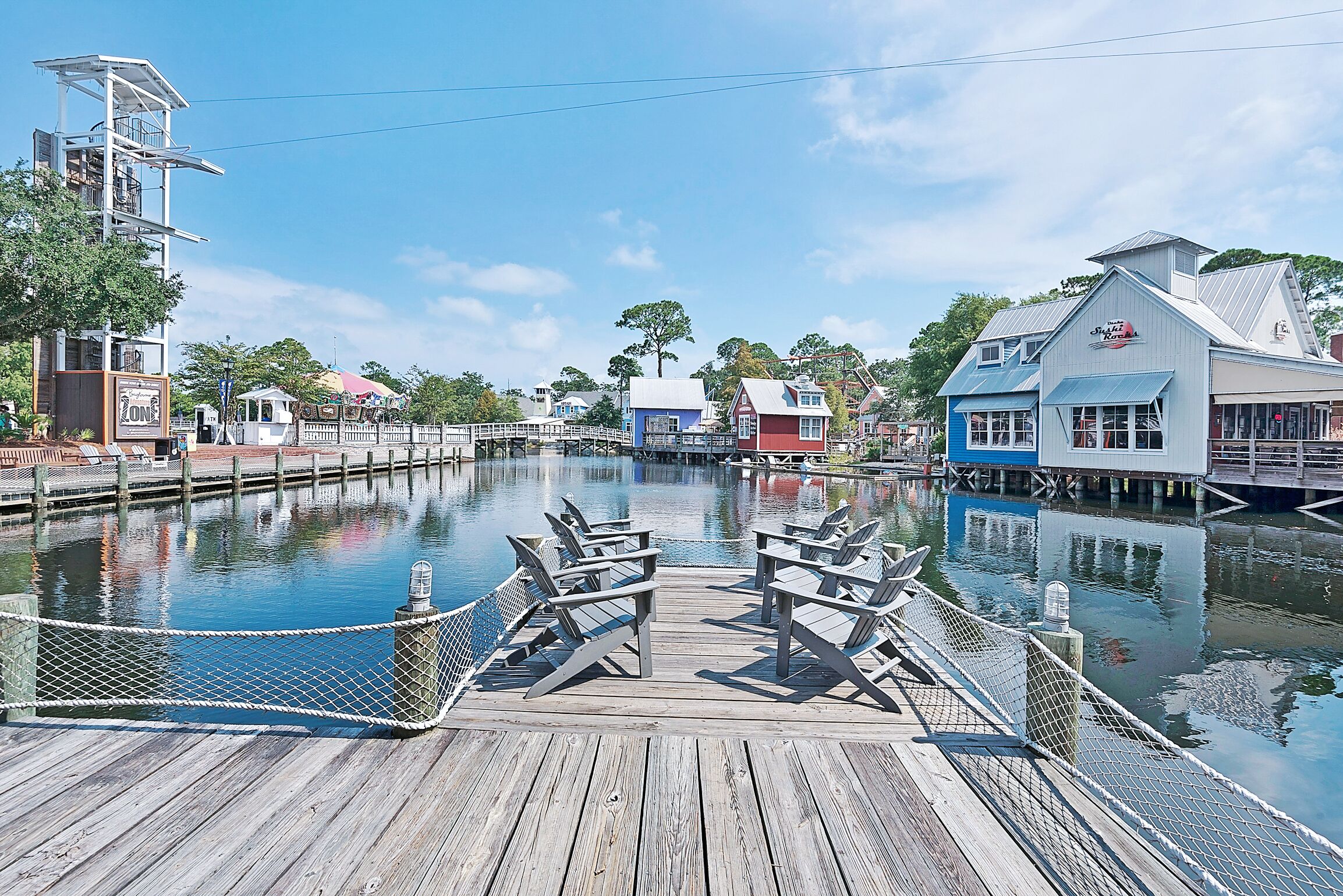 A wooden dock with chairs and a rope barrier over a calm water body, surrounded by buildings with distinct architectural styles.