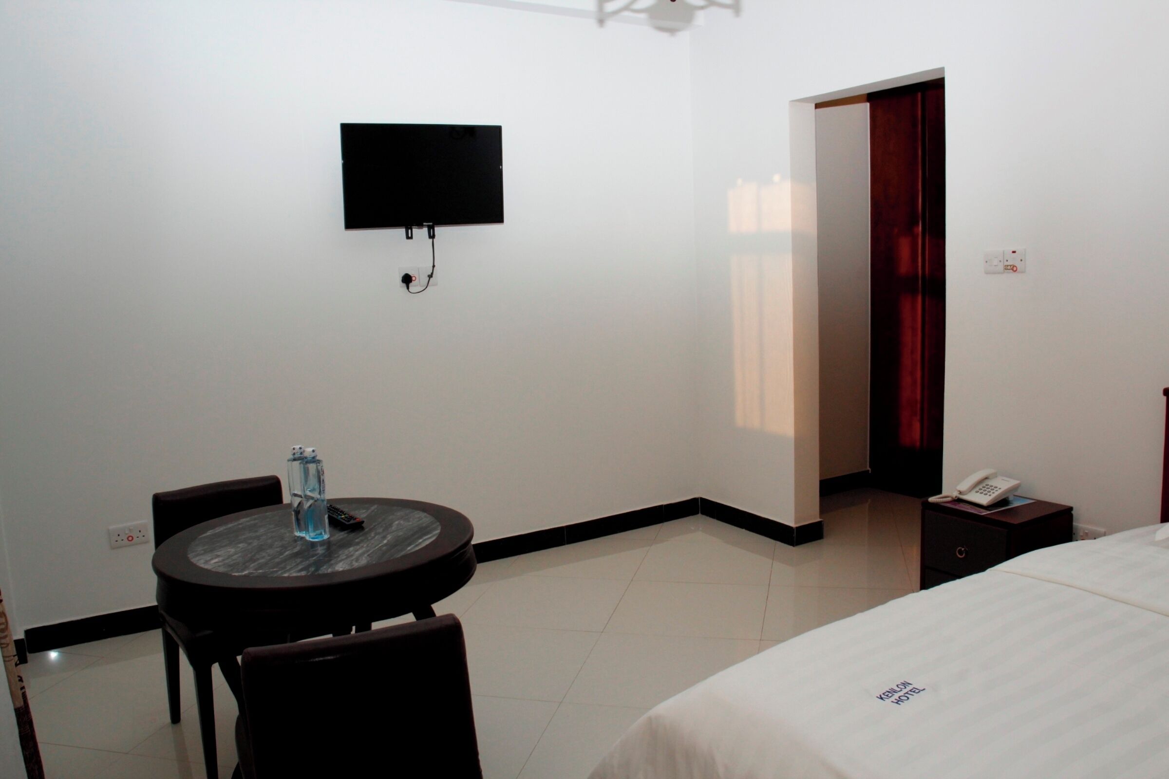 Quarto de hotel com cama, televisão na parede, pequena mesa com cadeiras e mesa de cabeceira com telefone.