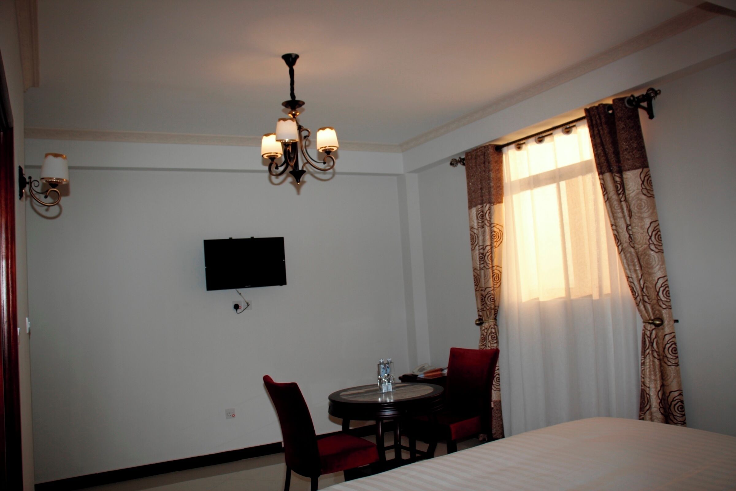 Ein Hotelzimmer mit Bett, Wandfernseher, rundem Esstisch mit Stühlen und einer Kristallleuchter.