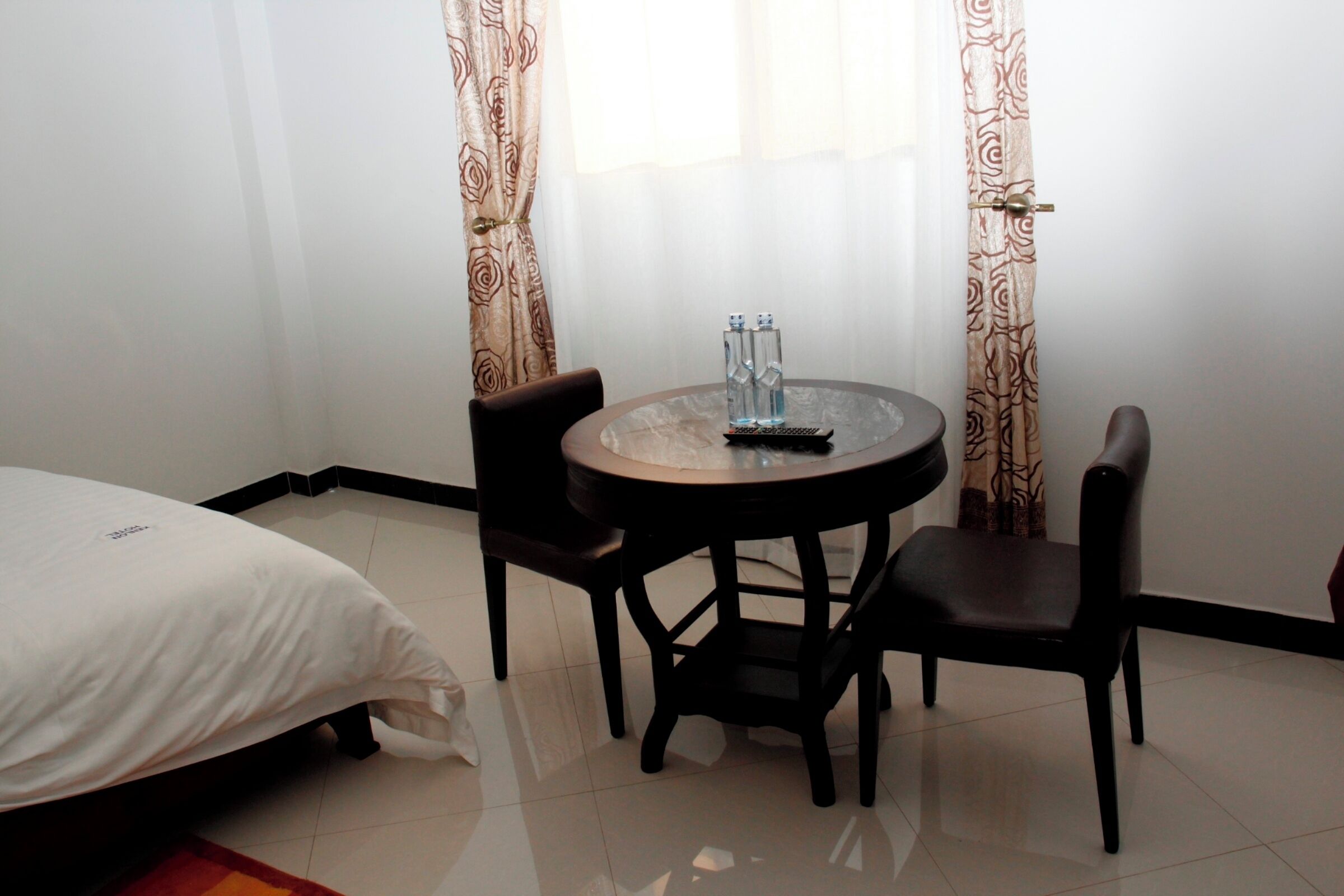 Una habitación de hotel con una cama, una mesa redonda de vidrio y dos sillas, y un ventanal con cortinas.