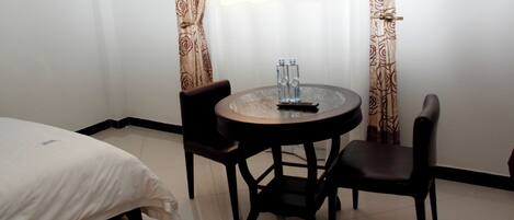 Una habitación de hotel con una cama, una mesa redonda de vidrio y dos sillas, y un ventanal con cortinas.