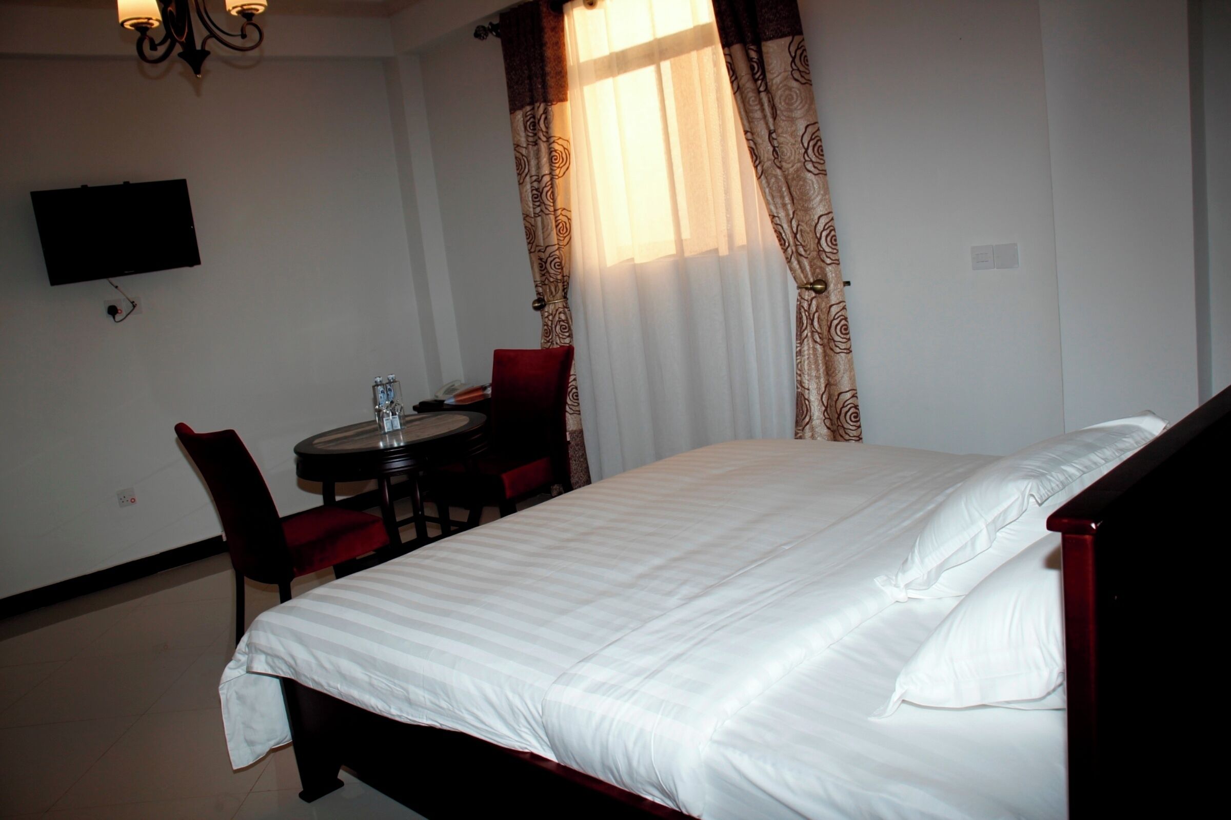 A hotel room with a bed, a TV mounted on the wall, a dining table with chairs, and a window with curtains.