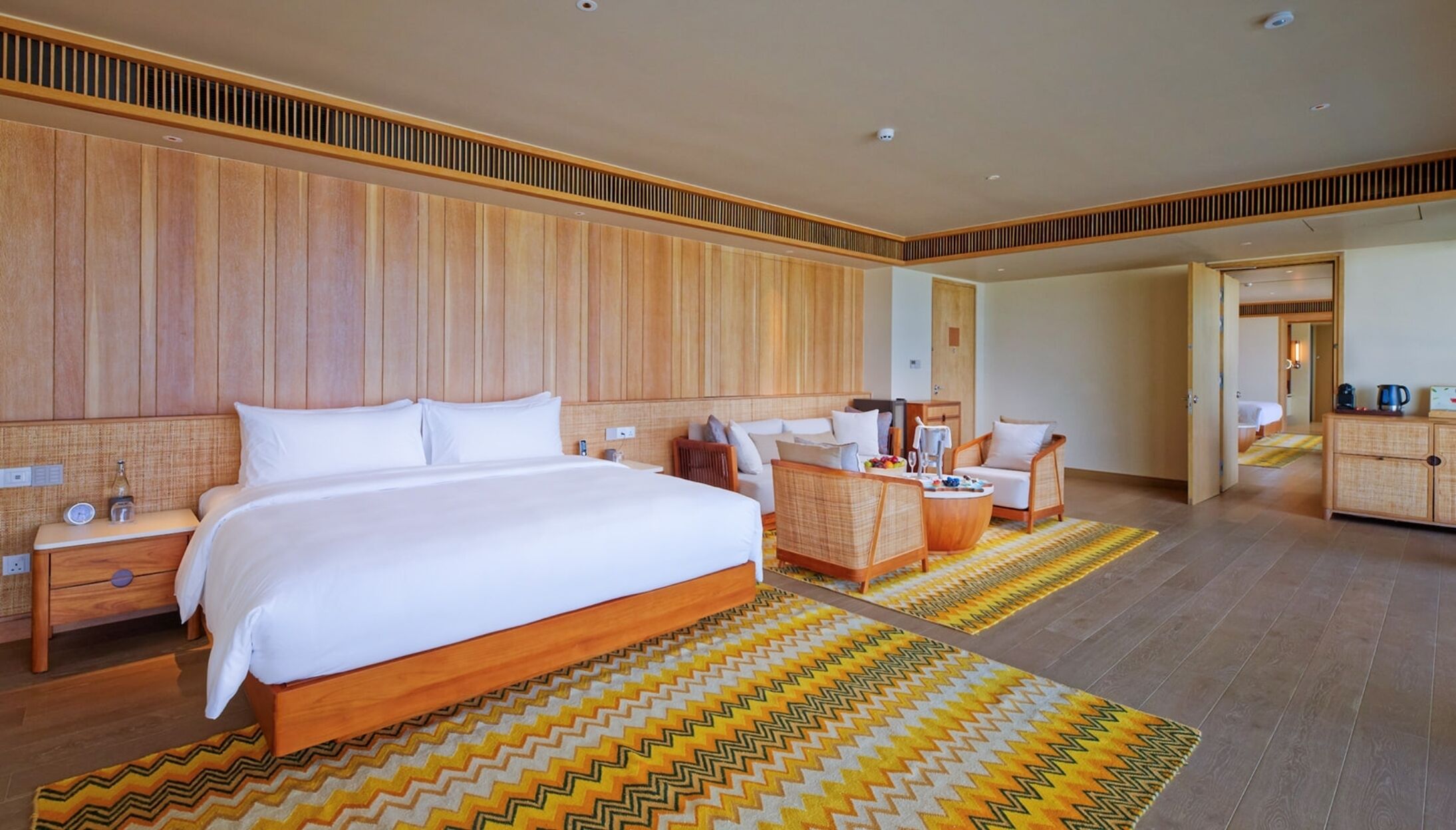 A hotel room with a large bed, a sitting area with a sofa and chairs, a wooden wardrobe, and a small table with a kettle.