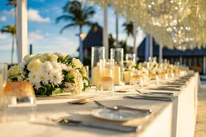 Ein langer Tisch, gedeckt mit weißen Blumen, Kerzen und klarem Glas, vermutlich für eine festliche Veranstaltung.