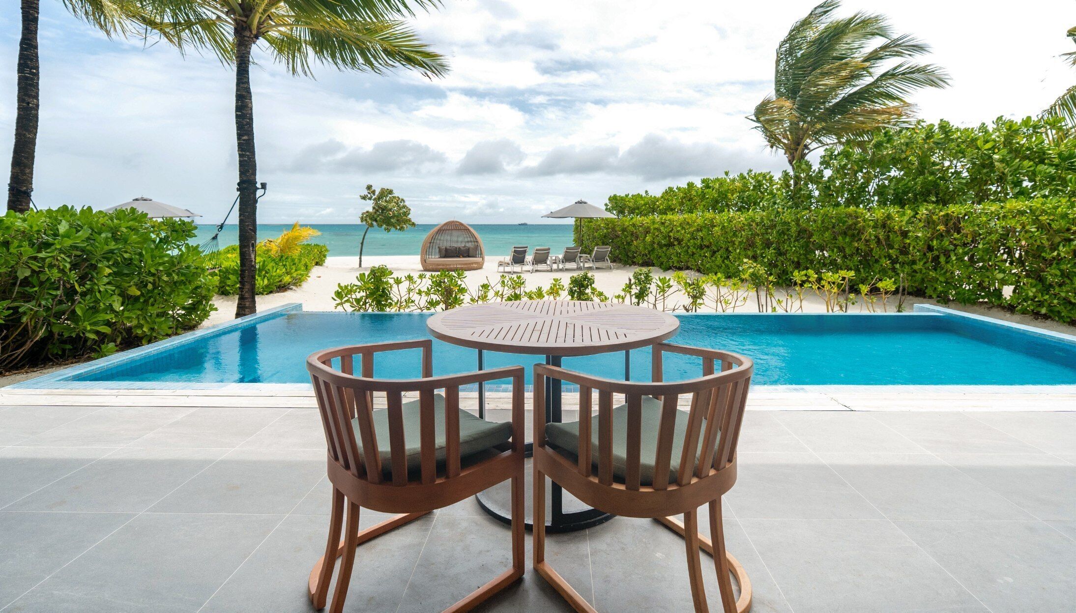 A poolside area with a round table and chairs, a view of the ocean, and a beach in the background.