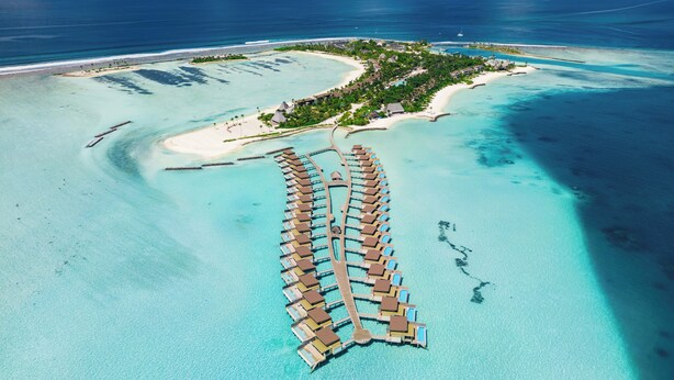 An aerial view of a resort with overwater bungalows connected by a wooden walkway, surrounded by turquoise waters and a small island with buildings and greenery. - Kuda Villingili Maldives, Free Return Transfers 03 nights & above (Kuda Villingili)