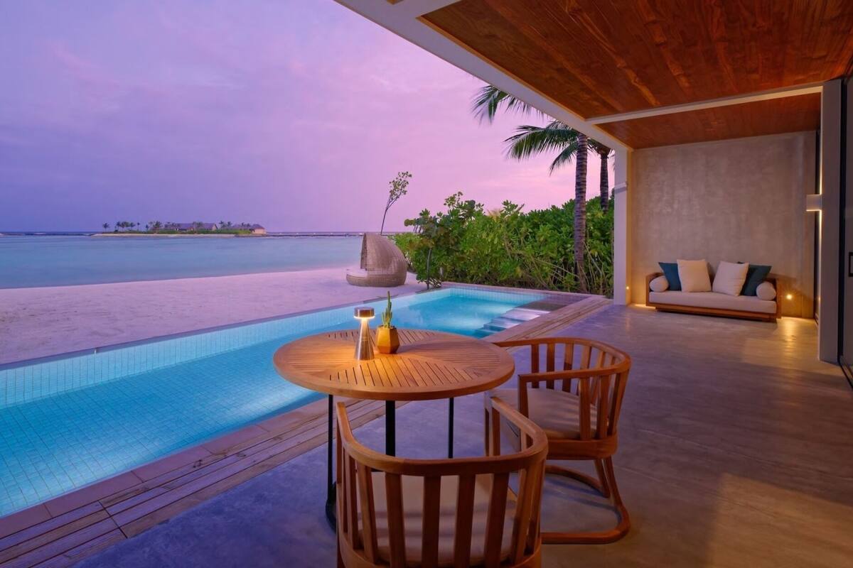 A poolside area with a wooden deck, a round wooden table with chairs, a lounge chair, and a sofa.