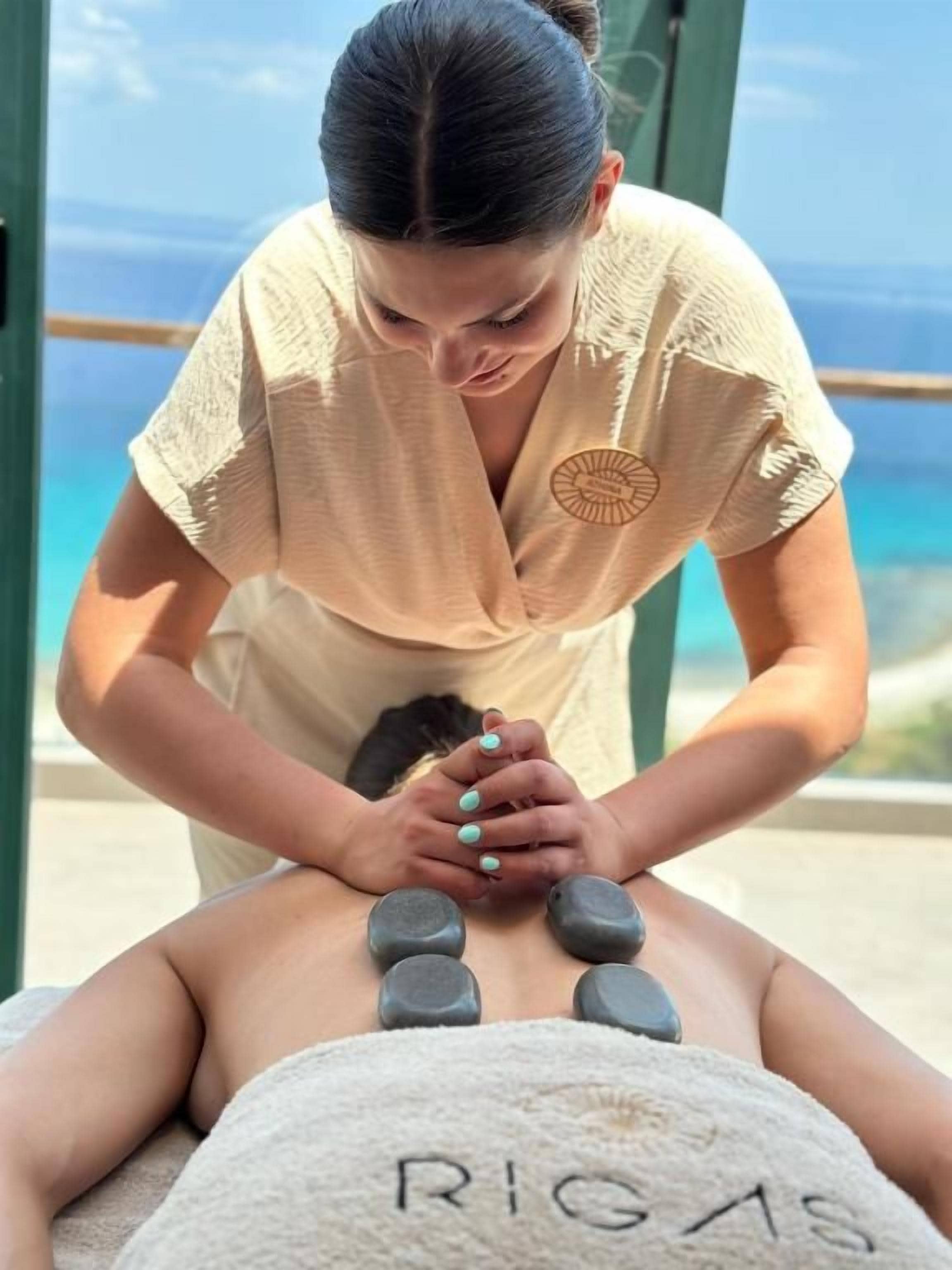Eine Massageanwendung: Eine Person liegt mit dem Gesicht nach unten, während eine Rückenmassage mit Steinen durchgeführt wird. Ein Handtuch mit der Aufschrift 'RIGAS' liegt ebenfalls bereit.