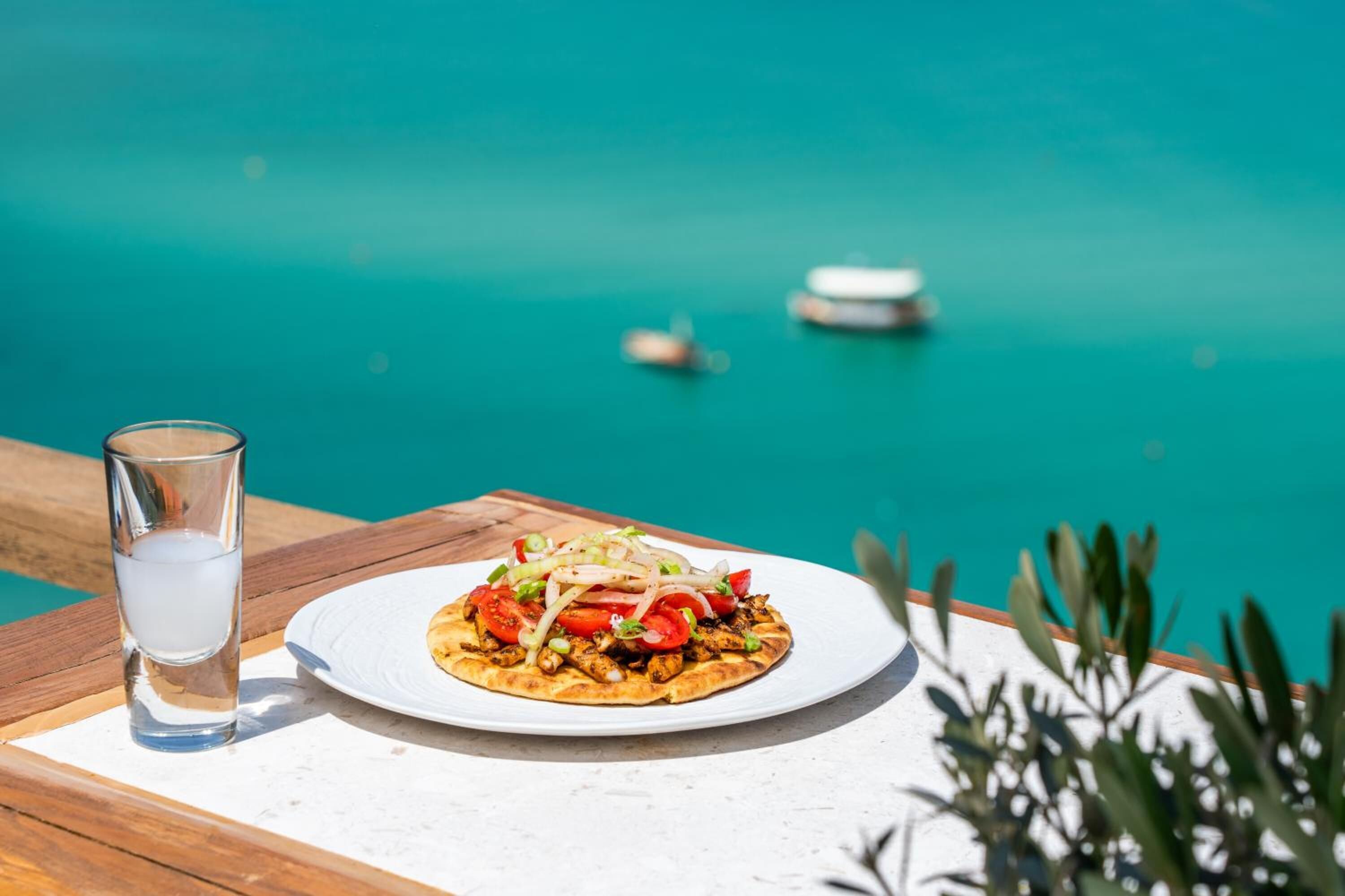 A plate of food on a wooden table with a glass of water beside it, overlooking a turquoise sea with boats.