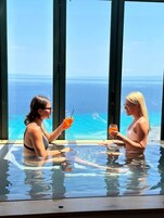 Dos personas en una piscina con fondo de cristal y vista al océano.