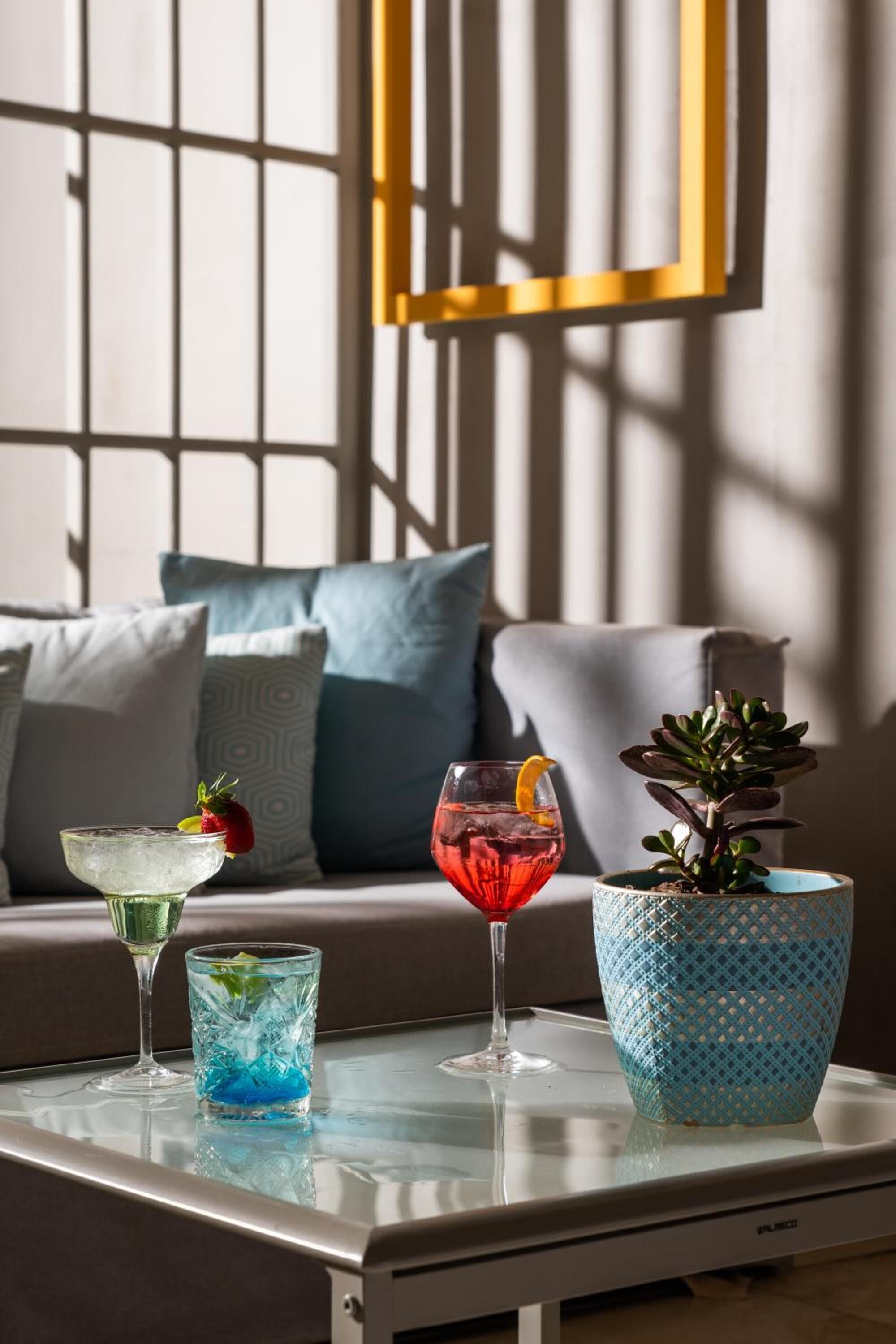 A glass table with three drinks, a sofa, and a potted plant in the background.