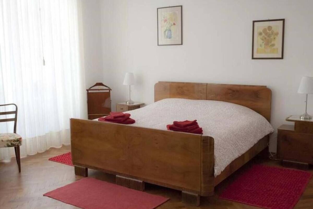 A bedroom with a wooden bed, two bedside tables, a chair, and a window with sheer curtains.