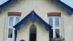 Un bâtiment à deux étages, doté d’un toit à pignon, de deux fenêtres à cadre blanc et d’une petite fenêtre en arc de cercle. On peut voir une plaque sur le mur et une enseigne indiquant « CHAPEL COTTAGE ».