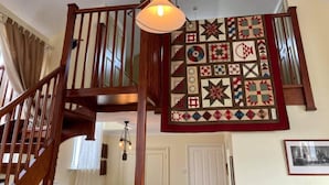 Un escalier avec une rampe en bois, un luminaire suspendu et une grande courtepointe accrochée au mur.