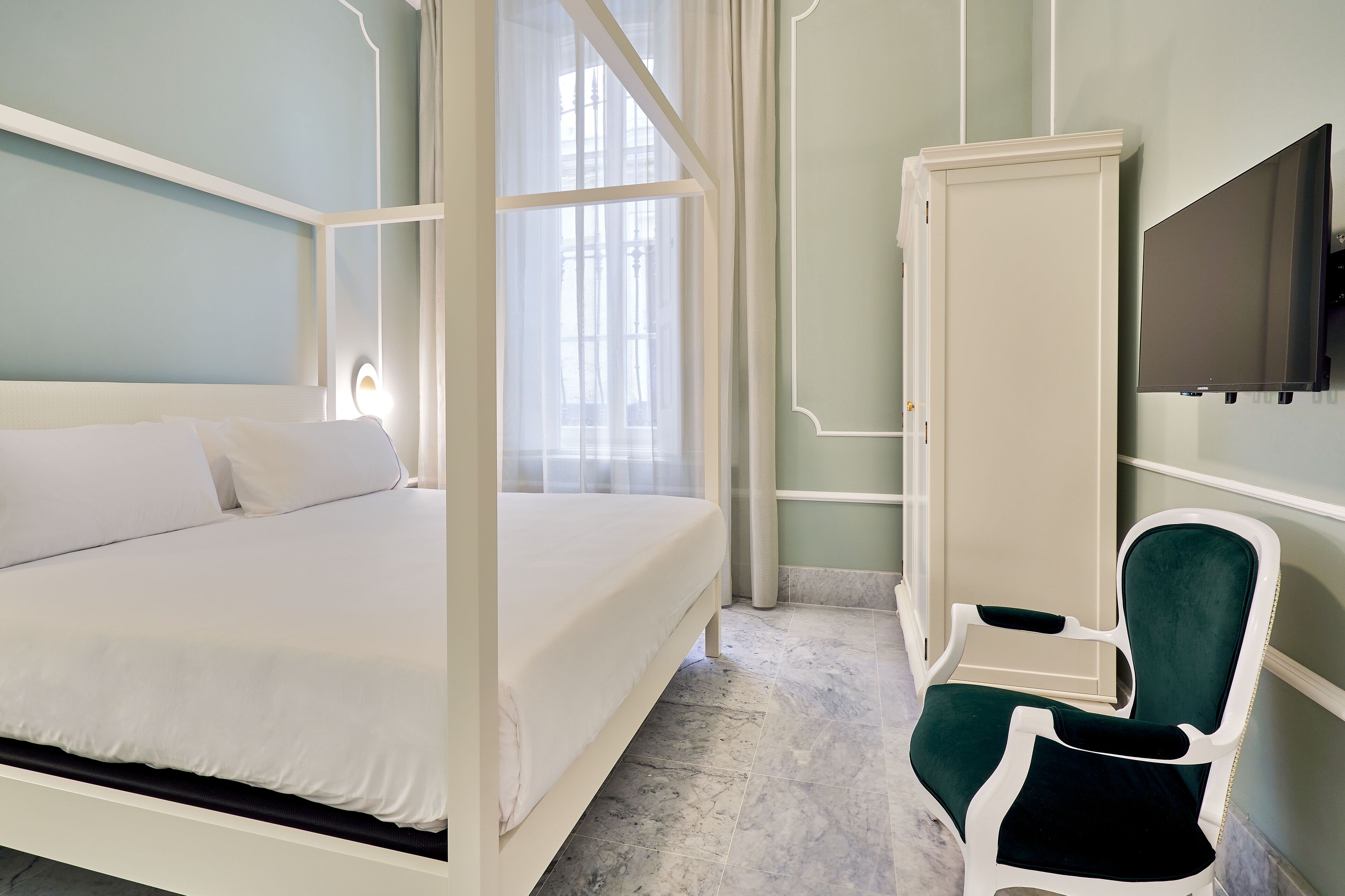 Un dormitorio con cama con dosel, una silla tapizada de verde, un televisor y un armario.