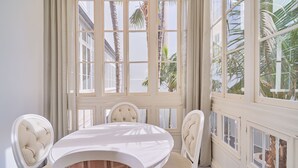 Uma sala de jantar com uma mesa redonda e quatro cadeiras, janelas amplas com vista para palmeiras e uma parede branca.
