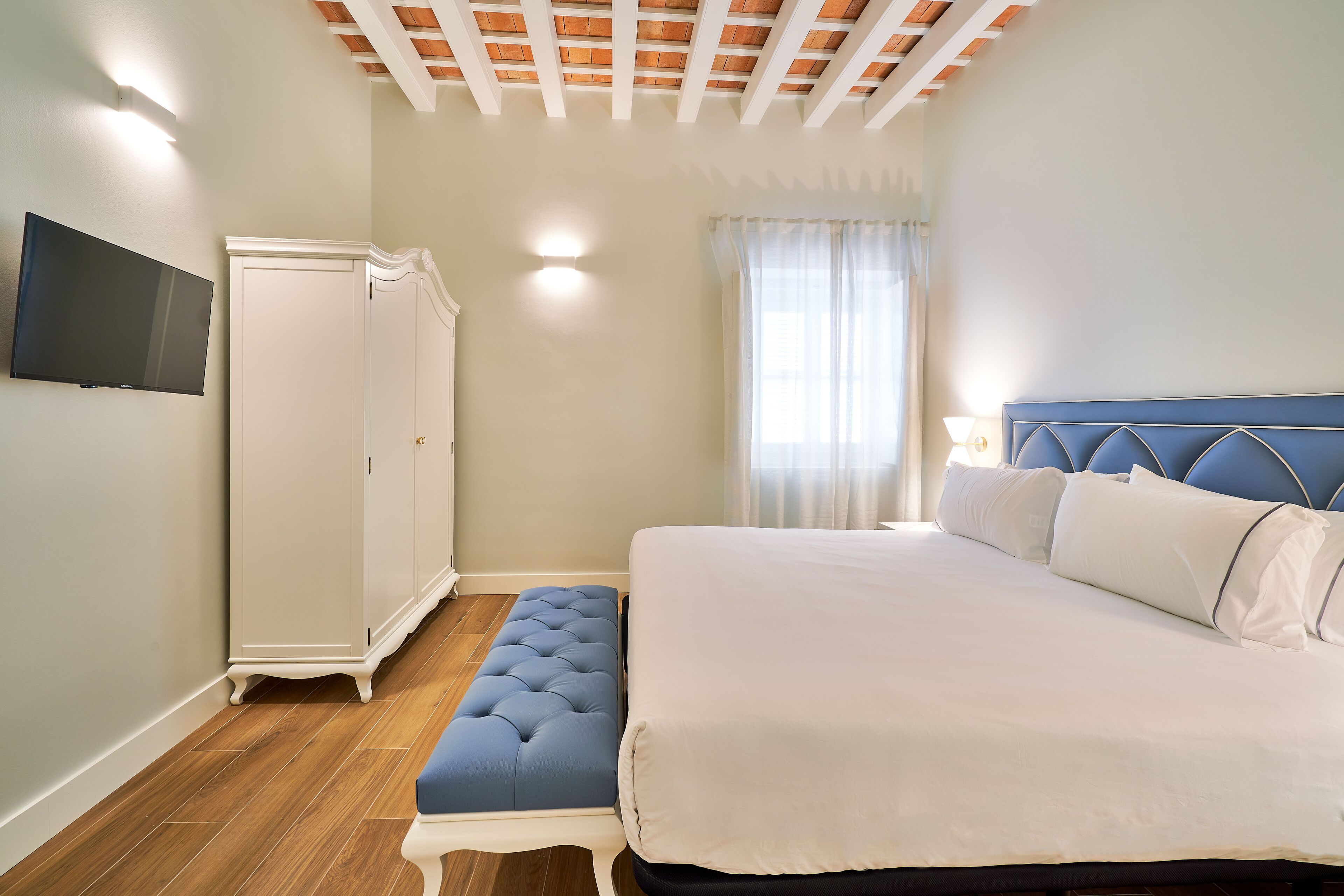 Een slaapkamer met een bed, een bankje, een kledingkast en een televisie aan de muur.
