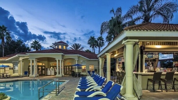 Ein Poolbereich mit Liegestühlen, einer Bar und einem Gebäude mit Säulen und einem Ziegeldach.