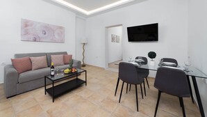 A modern living room with a sofa, coffee table, dining table, chairs, a flat-screen TV, and artwork on the walls.