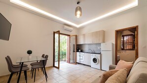 A modern kitchen with a built-in oven and a washing machine, a dining area with a glass table and chairs, and a living room with a sofa.