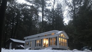 A cabin with a large front window, surrounded by snow-covered ground and tall trees. - Park Model Single Family double lofted private heaven on a pond (Otis)