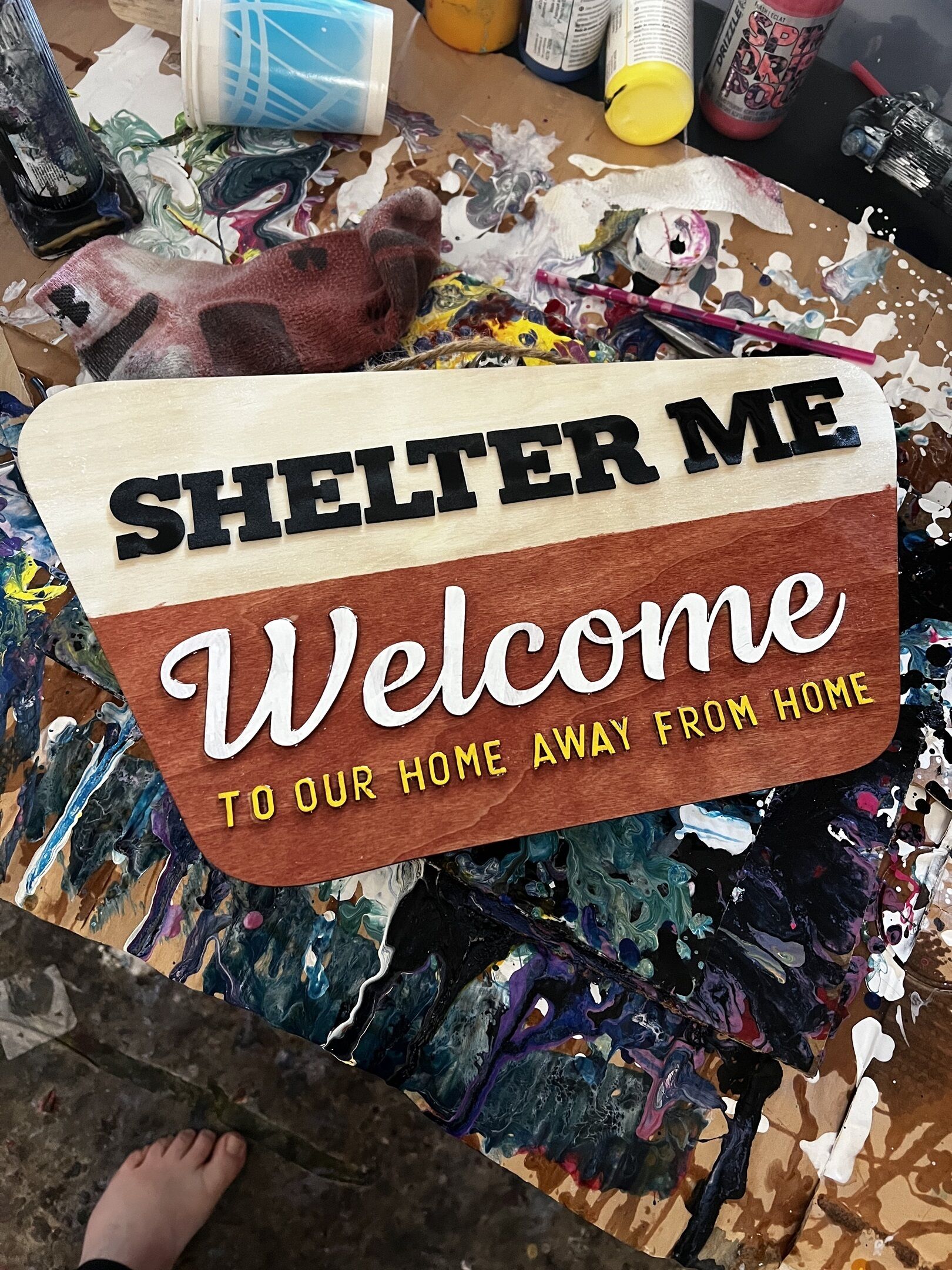 En velkomstskilt med teksten "SHELTER ME Welcome TO OUR HOME AWAY FROM HOME" mod en træbaggrund.