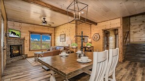 L’intérieur chaleureux d’un chalet avec une table  à manger en bois, des chaises blanches, une cheminée, un ventilateur de plafond et une grande fenêtre donnant sur le paysage extérieur.
