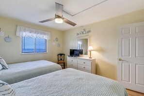 Une chambre avec deux lits, un ventilateur de plafond, une fenêtre avec des stores, une commode, un téléviseur et une porte.