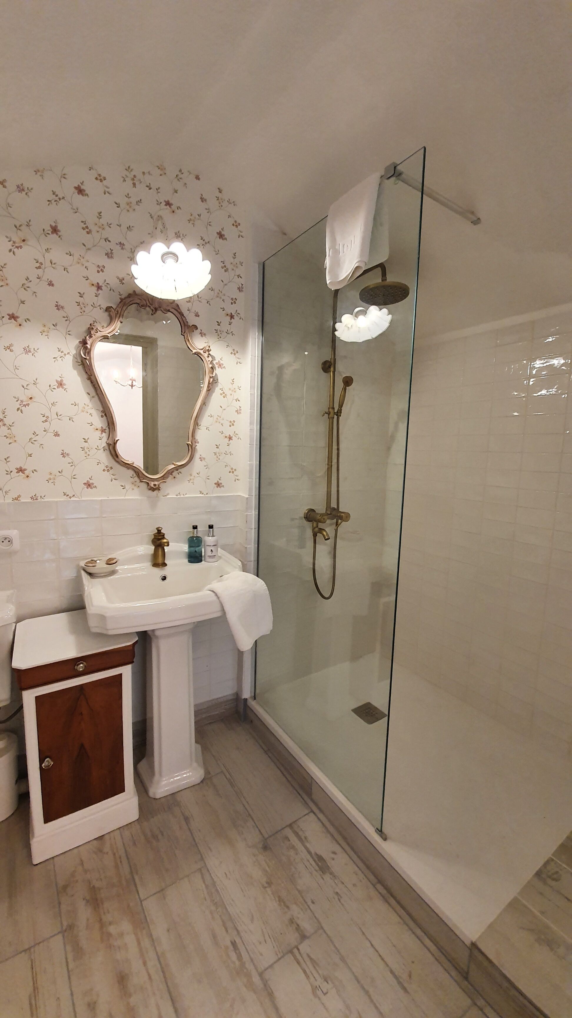 A bathroom with a glass-enclosed shower, a pedestal sink, and a mirror with a floral wallpaper.