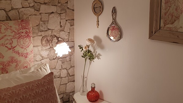 A bedroom with a stone wall, a bed with floral bedding, a bedside table with a vase of flowers, and wall-mounted mirrors. - Hideaway gite for 2 with private garden and sundeck, close to Atlantic beaches. (Épargnes)