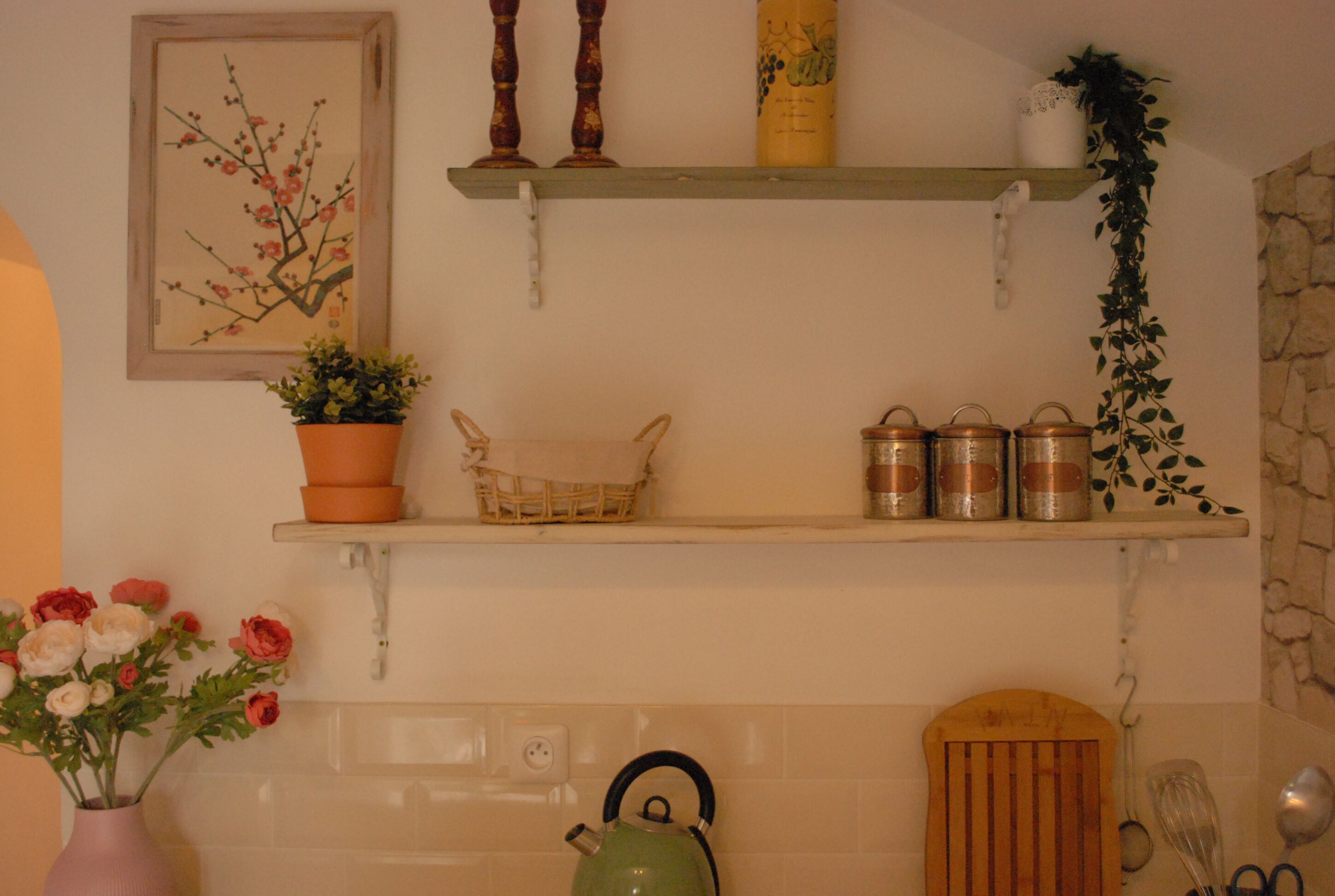 Une cuisine avec une bouilloire verte, un vase de fleurs et des étagères remplies de bocaux et d’une plante.