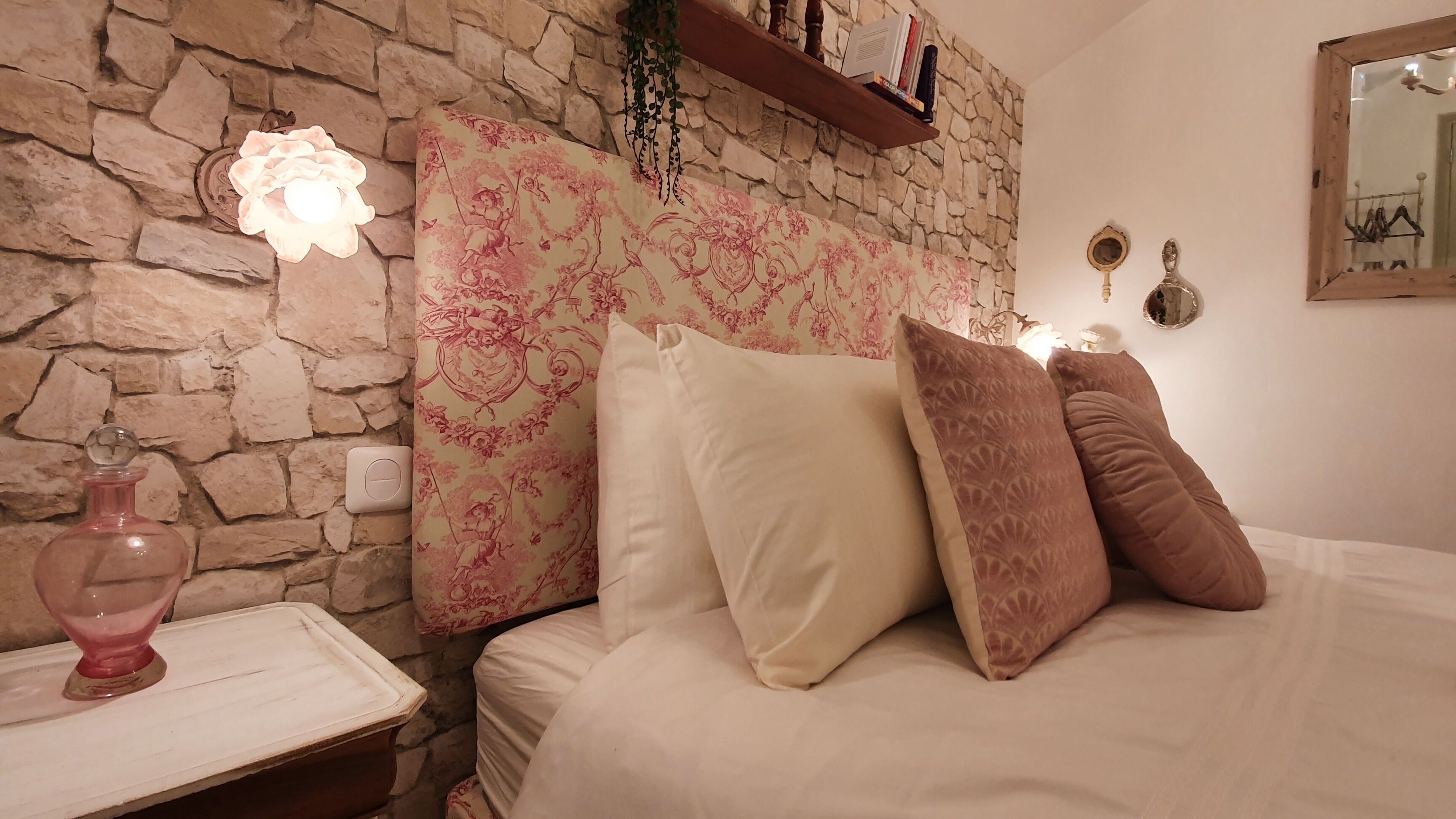 Un dormitorio con pared de piedra, una cama con almohadas blancas y marrones, una mesita de noche con un objeto rosa, y una estantería con libros y un espejo.