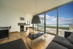 A modern living room with a fireplace, a dining area, and a large window offering a view of the sea.