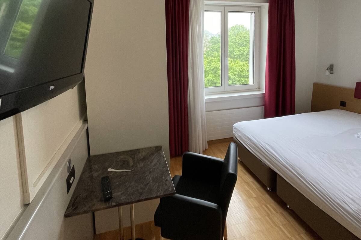 A hotel room with a bed, a desk with a chair, a TV mounted on the wall, and a window with curtains.