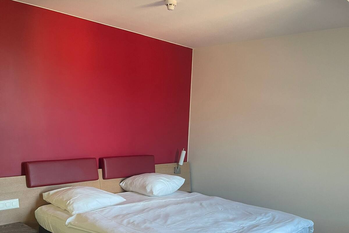 A bedroom with a bed, wooden flooring, a red accent wall, and a ceiling light.