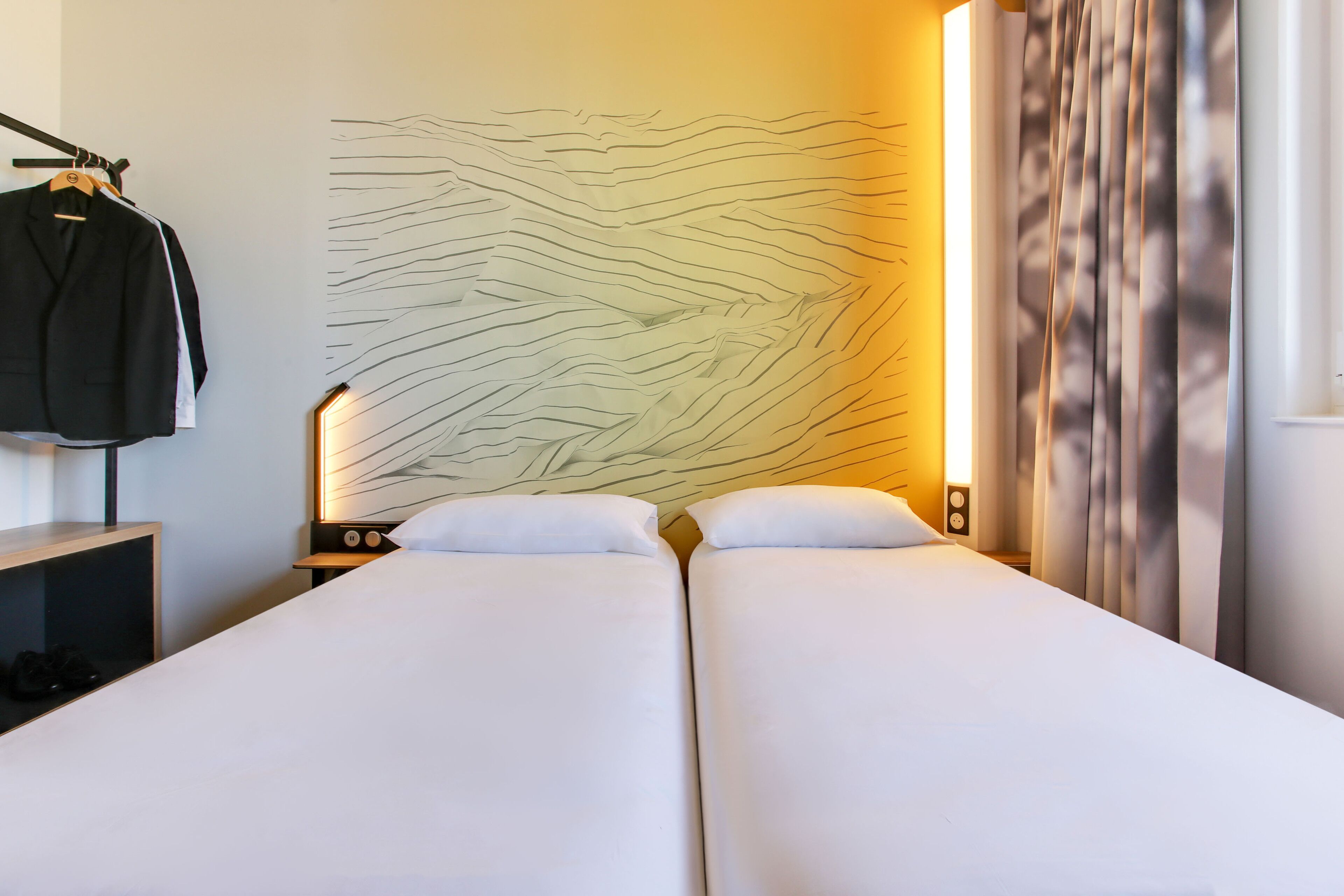 A hotel room with two beds, a coat rack, a wall art piece, and a curtain.