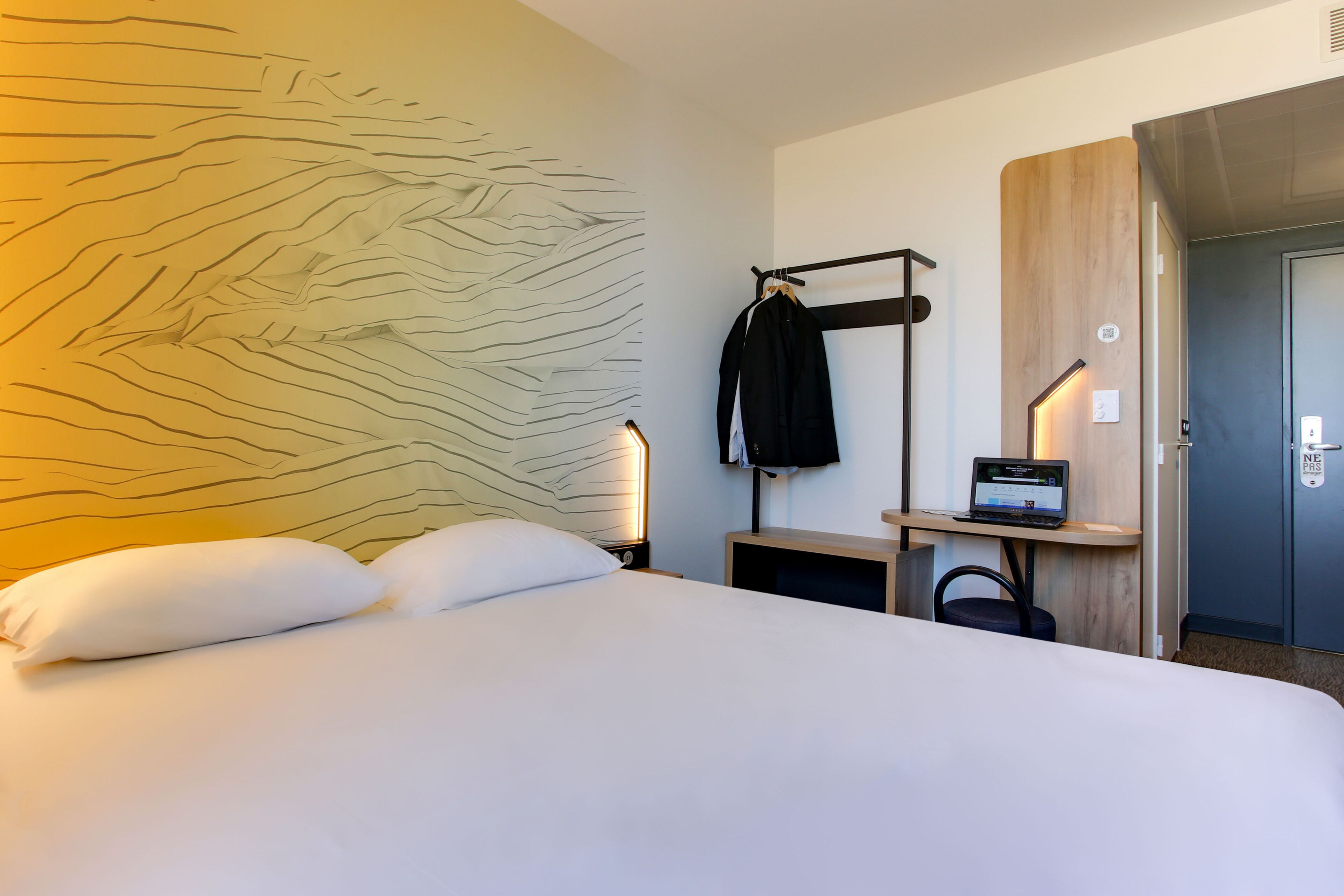 Habitación de hotel con una cama grande, perchero en la pared, escritorio con una computadora portátil y una puerta con un cartel que dice 'NE PAS'.