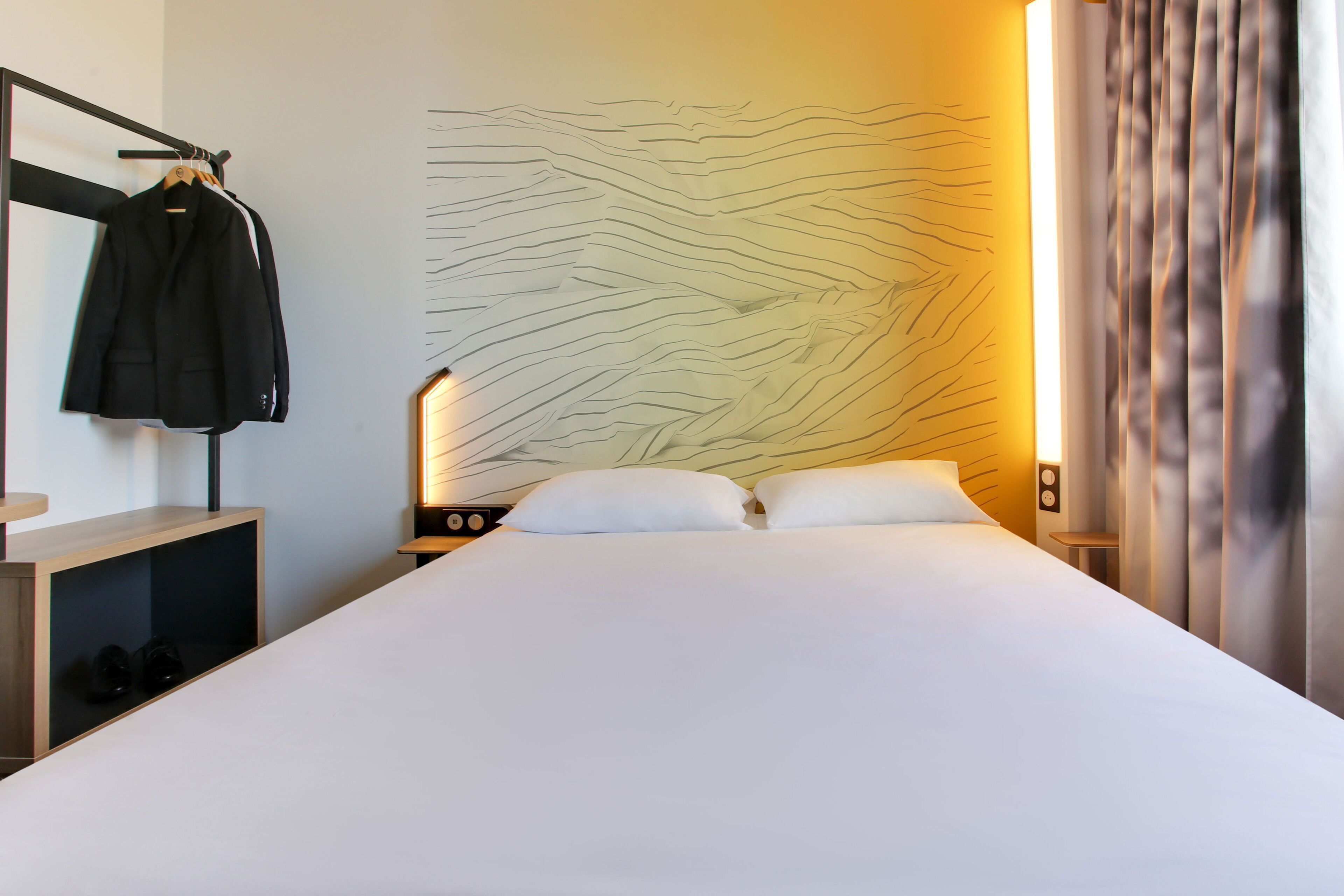 A hotel room with a large bed, a coat rack with a jacket, a wall with a wavy pattern, and a nightstand with a lamp.