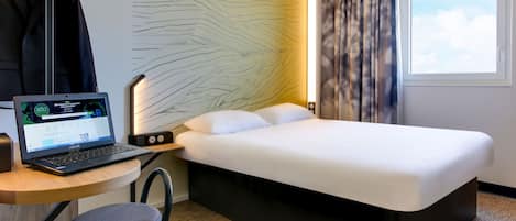 A hotel room with a bed, a desk with a laptop, a hanging coat, and a window with curtains.