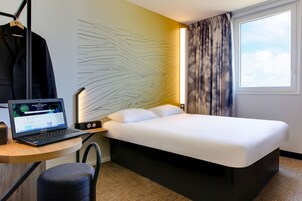 Una camera d'albergo con un letto, una scrivania con un portatile, un cappotto appeso e una finestra con tende.
