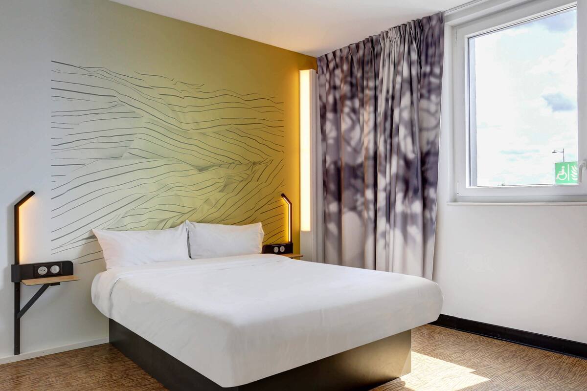 A modern hotel room with a large bed, a headboard with a unique light design, a nightstand with a lamp, and a view of the city through the window.