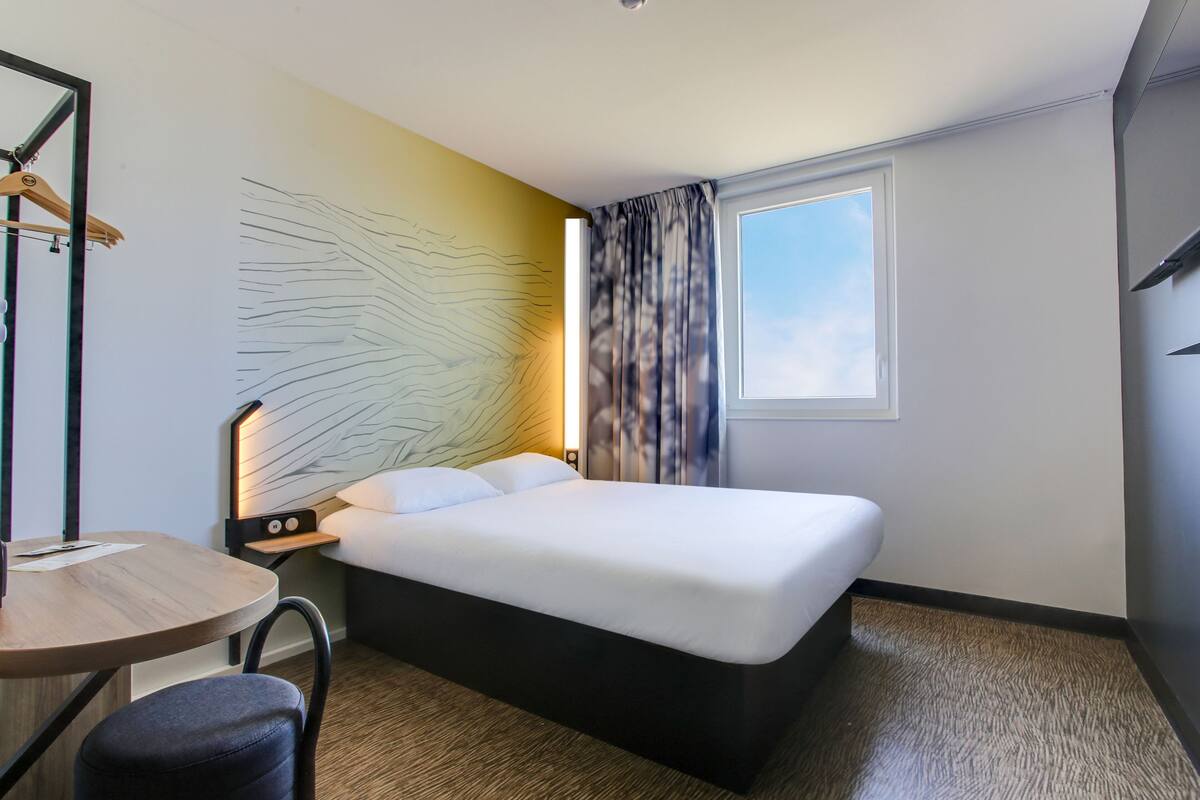 A hotel room with a bed, a desk, a chair, a wall art piece, and a window with curtains.