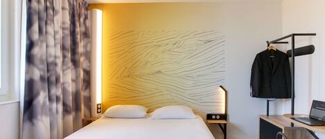 A hotel room with a bed, a desk with a laptop, a chair, a coat rack, and a wall with a light fixture.
