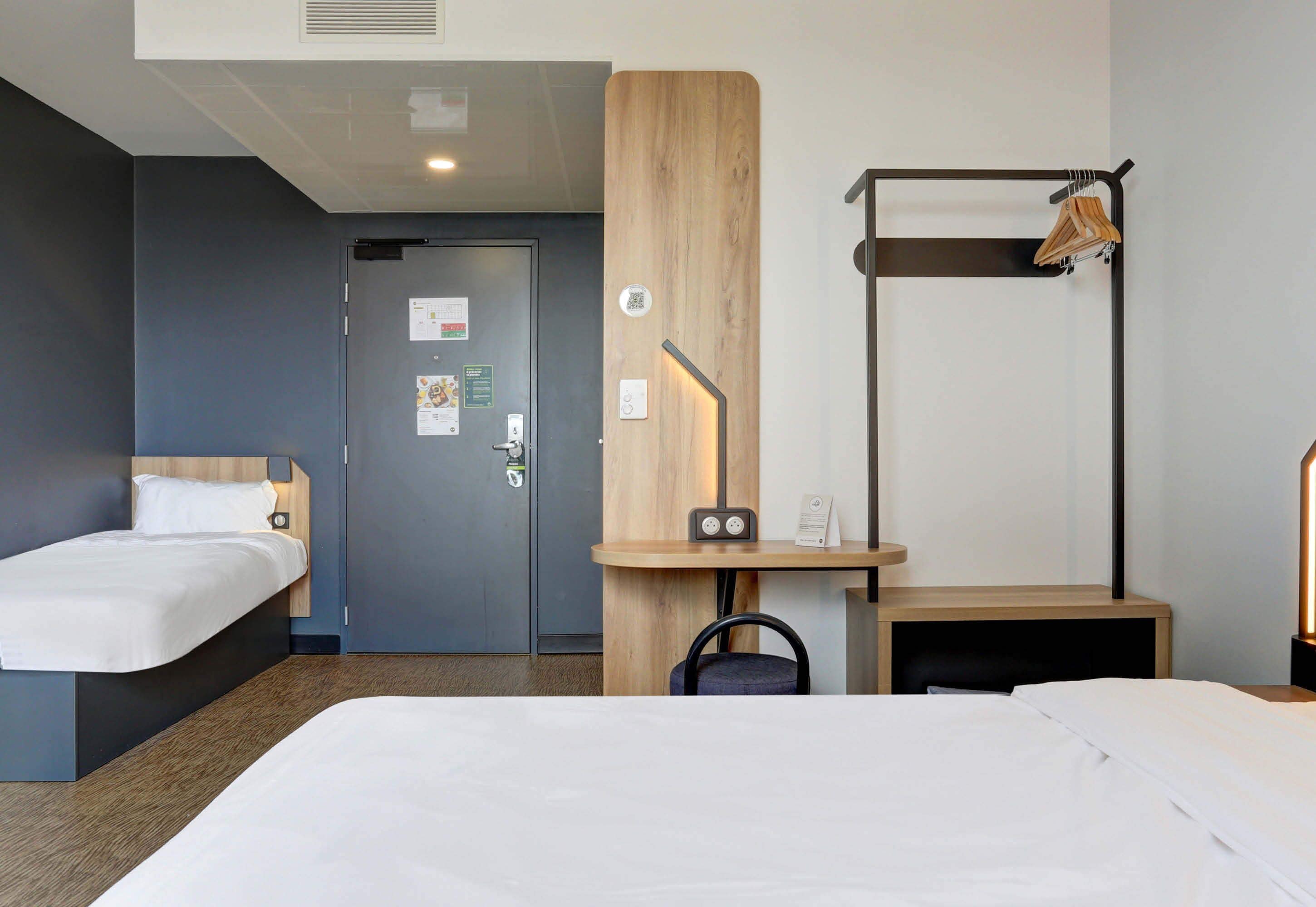 A hotel room with two beds, a wooden accent wall, a desk with a chair, a coat rack, and a door with a QR code.