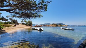 Eine klare Küstenlandschaft mit in ruhigem Wasser liegenden Booten, einem Sandstrand und einer felsigen Landzunge mit Vegetation.