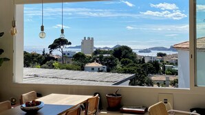 Una habitación con un ventanal grande con vistas a un pueblo costero, un comedor con mesa y sillas, y lámparas colgantes.