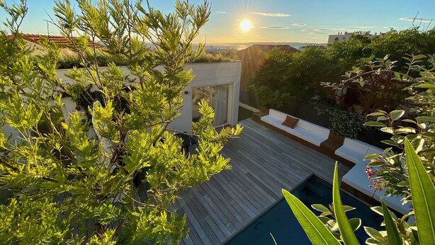 Un área de piscina en la azotea con terraza de madera, zona de estar exterior blanca y abundante vegetación.